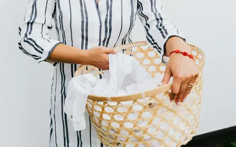 Manos sostienen una cesta de mimbre llena de ropa blanca, lista para lavar camiseta a mano.