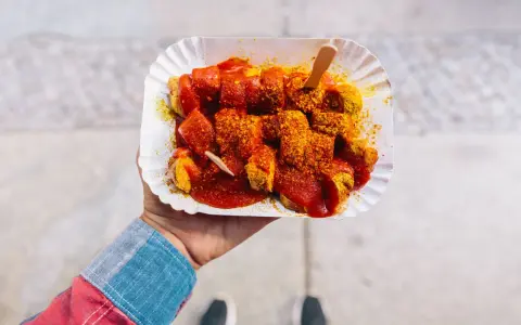 Eine Hand hält eine Portion Currywurst mit original Currywurst Sauce, bestreut mit Currypulver. Ein Holzspieß steckt darin.