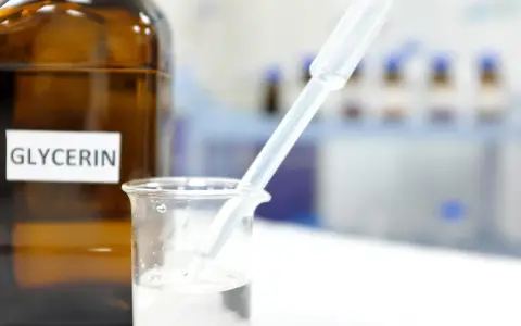 Braunglasflasche mit der Aufschrift "Glycerin". Ein Pipette tropft in ein Becherglas mit klarer Flüssigkeit, was die Glycerin verwendung in der Forschung zeigt.