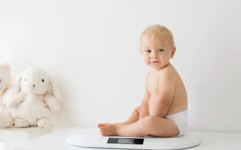 Uśmiechnięty chłopczyk w pieluszce siedzi na wadze. Chyba już czas na większy rozmiar, kiedy pampers jest za mały.