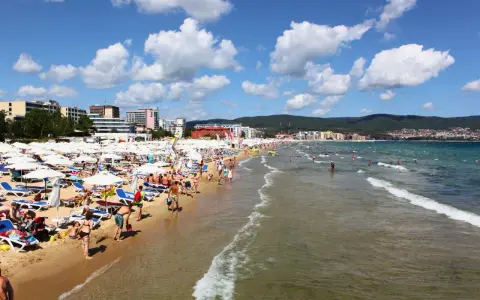 Tłum ludzi wypoczywa na piaszczystej plaży Słoneczny Brzeg, opalając się na leżakach pod białymi parasolami. W tle widać morze i zabudowania kurortu.