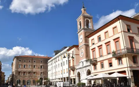 Piazza Tre Martiri a Rimini: un campanile con orologio domina la scena, tra palazzi storici e tavolini all'aperto. Ecco cosa vedere!