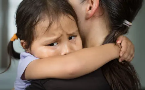 Ein Kind weint im Kindergarten und klammert sich an seine Mutter. Die Kleine hat Tränen in den Augen und sucht Trost.