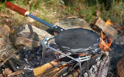 En gjutjärnspanna med trähandtag står på en ställning över en öppen eld. Perfekt för att steka plättar eller våfflor ute i naturen.