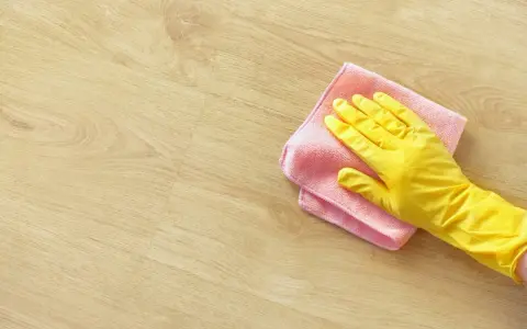 En hand i gul handske torkar av ett ljust trägolv med en rosa trasa. Perfekt för pigmenterad olja till ekbord.