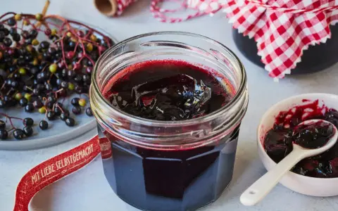 Frisch zubereitete holunderbeeren marmelade im Glas, daneben frische Beeren und eine kleine Schale mit Löffel.