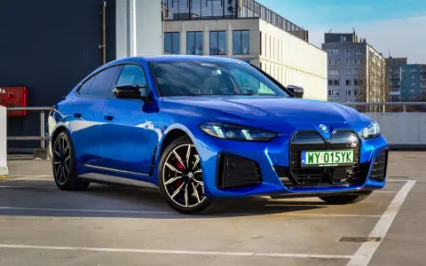 Niebieskie BMW i4 M50 zaparkowane na parkingu, z widokiem na budynki w tle.