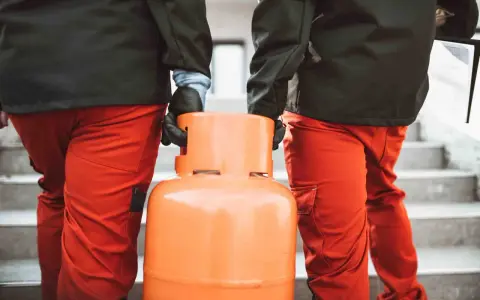 Dwóch mężczyzn w pomarańczowych spodniach niesie butlę z gazem. Wygląda na to, że nie można zakręcić butli z gazem, co może być problemem.