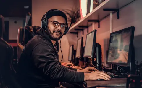 Młody game developer w słuchawkach i okularach pracuje przy komputerze w ciemnym pomieszczeniu.