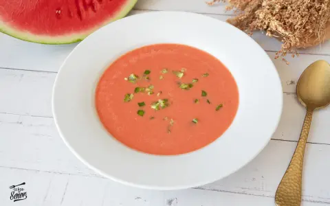Un refrescante gazpacho de sandía, adornado con trocitos verdes, listo para disfrutar junto a una rodaja de sandía y una cuchara dorada.