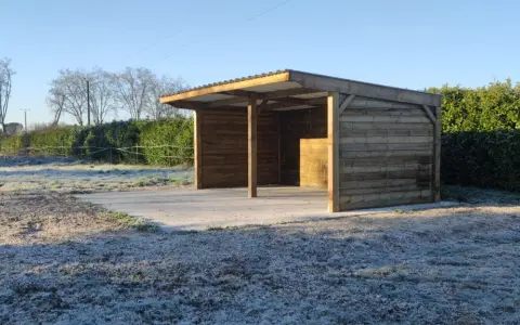 Abri pour chevaux d'occasion en bois, avec toit métallique, sur un sol gelé. Parfait pour protéger vos équidés du froid.