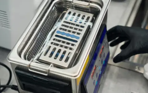Ein Handschuh bedient ein Gerät, das zeigt, wie funktioniert ein ultraschallreiniger. Kleine Instrumente liegen in einem Korb im Wasserbad.