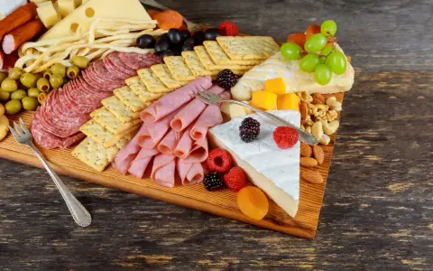 Apetyczna deska serów i wędlin: salami, szynka, ser żółty, pleśniowy, oliwki, winogrona, maliny, orzechy i krakersy.