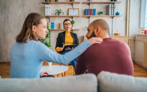 Un couple en consultation avec une psychologue conjugale. La femme pose une main réconfortante sur l'épaule de son partenaire.