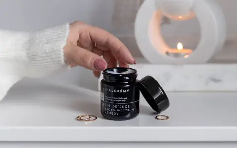 D'ALCHEMY - krem do twarzy, szyi i dekoltu. Dłoń sięga po słoiczek, w tle pali się świeca.