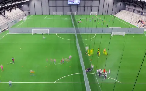 Barn tränar fotboll på en grön konstgräsplan i Prioritet Serneke Arena. Publik sitter på läktaren.