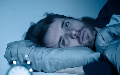 Mann liegt wach im Bett, Augen weit offen, die Angst vor Schlaflosigkeit ist ihm ins Gesicht geschrieben. Ein Wecker ist im Vordergrund zu sehen.