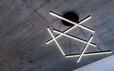 Oryginalna lampa sufitowa z geometrycznymi, podświetlanymi ramionami, zawieszona na tle surowego, betonowego stropu.