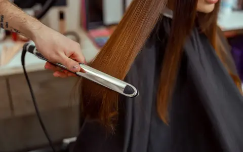 Stylista pokazuje, jak zrobić fale prostownicą, tworząc delikatne loki na długich, brązowych włosach.