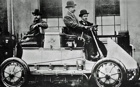 Hombres con sombreros posan en el primer coche eléctrico de la historia, un vehículo pionero de finales del siglo XIX.