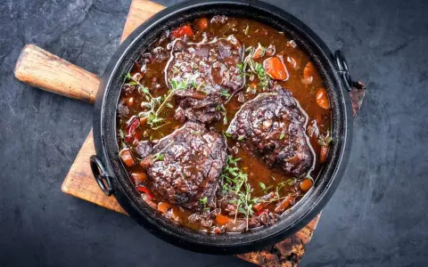 Carrilleras de ternera estofadas en salsa oscura con zanahorias y hierbas, servidas en una cazuela de hierro fundido sobre tabla de madera.