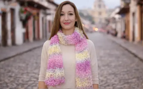Mujer sonriente luce bufandas de dos agujas modernas en tonos pastel, paseando por una calle empedrada.
