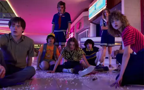 Los personajes de Stranger Things, con Eleven al frente, sentados en el suelo de un centro comercial, evocando las icónicas canciones de la serie.