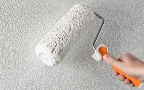 Eine Hand mit orangefarbenem Griff hält eine Farbrolle, die weißen Putz auf eine strukturierte Wand aufträgt. Der Anschluss Putz an Holz wird hier perfektioniert.
