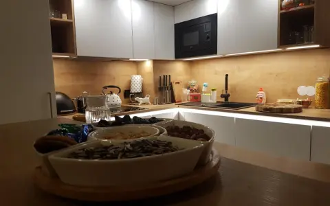 Nowoczesna kuchnia z białymi frontami i drewnianym blatem. Na blacie czajnik, zlew i naczynia z bakaliami. W tle wbudowany czarny okap, który idealnie komponuje się z całością, pokazując jak obudować komin w kuchni.