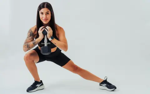 Donna con tatuaggi esegue affondo laterale con manubrio, dimostrando cos'è la palestra funzionale.