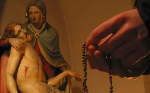 Eine Hand hält einen Rosenkranz, während die Pietà-Statue im Hintergrund zu sehen ist. Ein evangelischer Rosenkranz wird hier in Andacht gehalten.