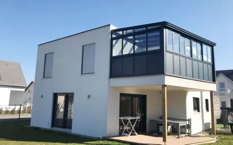 Maison moderne avec extension étage sur piloti. Une véranda vitrée surplombe une terrasse en bois.
