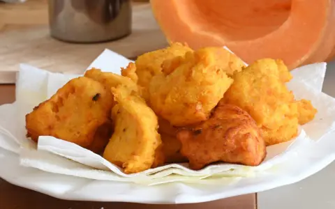 Frittelle di zucca dorate e croccanti, servite su carta assorbente, con una fetta di zucca sullo sfondo.