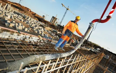 Un ouvrier en tenue orange et bleue coule du béton pour un mur banché sur un chantier. Grues et structures en construction en arrière-plan.