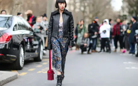 Frau im typisch 90er Grunge Style mit Lederjacke, geblümtem Kleid und Stiefeln. Ein roter Nietenbeutel rundet den Look ab.