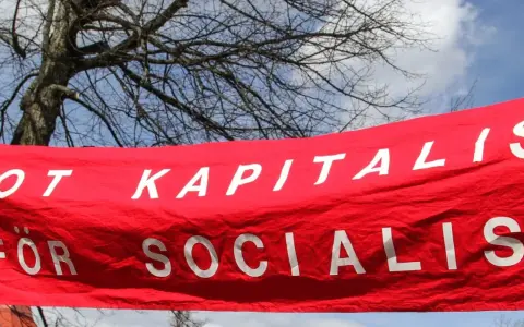 Röd banderoll med vit text: "MOT KAPITALIS FÖR SOCIALIS". En demonstration som visar vad socialism betyder.