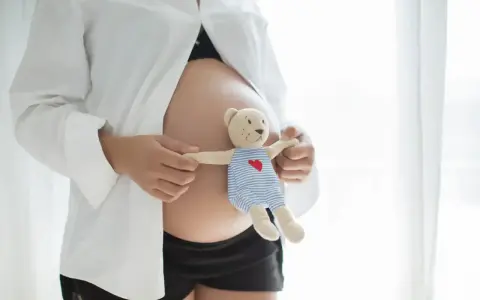 Brzuszek w 21 tygodniu ciąży, otulony białą koszulą. Dłonie trzymają misia w pasiastym ubranku, symbolizującego przyszłe macierzyństwo.