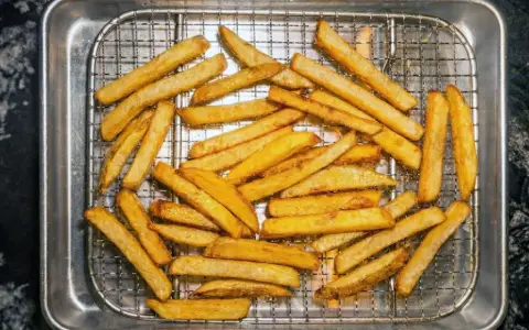 En hög med gyllene, krispiga pommes frites ligger på ett galler i en plåt. Perfekt för en snabb fika!
