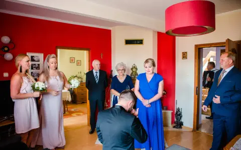 Nowoczesne błogosławieństwo: pan młody klęka przed panną młodą, otoczeni przez rodzinę i świadków.