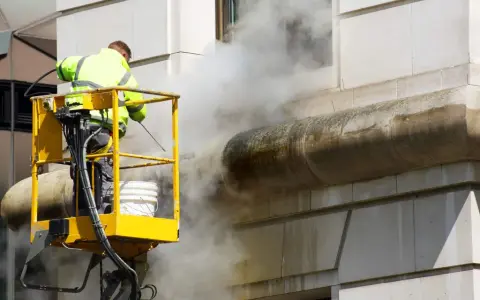 Un ouvrier en nacelle jaune utilise un nettoyeur haute pression pour décaper la peinture d'une façade en pierre, créant un nuage de vapeur.