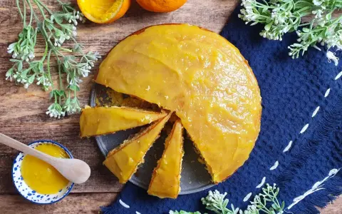 Una torta all'arancia morbidissima, con glassa lucida e profumata, pronta per essere gustata. Accanto, una ciotolina con crema all'arancia e un cucchiaino di legno.