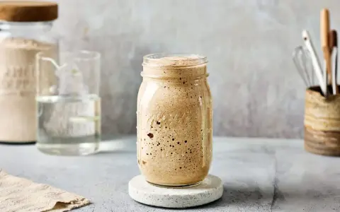 Ein Glas mit sprudelndem Sauerteig, angesetzt mit Dinkelmehl. Daneben ein Glas Wasser und ein Behälter mit Küchenutensilien.