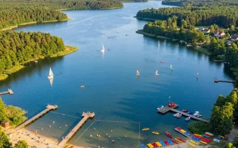 Letnie popołudnie nad jeziorem Kałębie. Żaglówki i kajaki na wodzie, plażowicze na piaszczystym brzegu, otoczonym zielonym lasem.