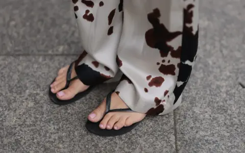 Delikatne, różowe paznokcie u stóp po zabiegu pedicure japoński, w czarnych klapkach, na tle spodni w cętki.