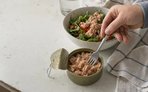 Una mano prende il tonno da una scatoletta aperta per aggiungerlo a un'insalata. Quanto tonno mangiare? Una porzione sana per un pasto leggero.