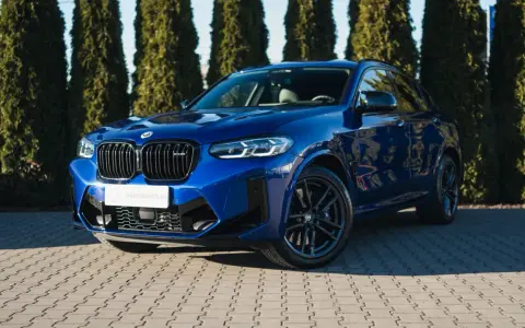 Błyszczący, niebieski suv BMW X4 M Competition zaparkowany przed żywopłotem.