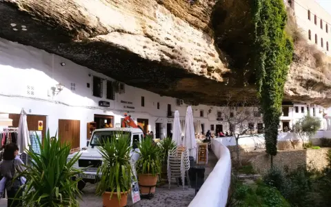 Häuser unter Felsvorsprung in Setenil de las Bodegas. Ein Auto und Pflanzen vor dem "El Gastor" Bar.