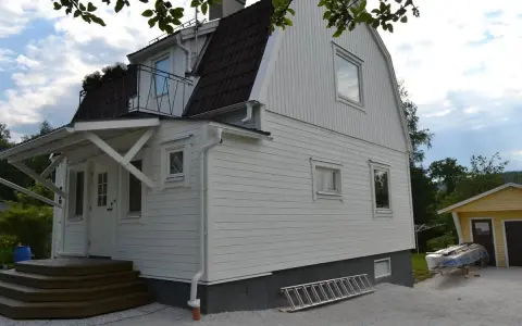Vitt hus med ny fasad, stegar och material för byta fasad. En gul garagebyggnad syns i bakgrunden.