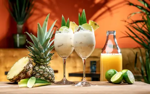 Två krämiga drinkar med ananasjuice, is och lime. En hel och en halv ananas samt en flaska juice står bredvid.