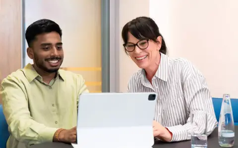 Ein junger Mann und eine Frau lachen über den Bildschirm eines Tablets. Sie sind bei einem Praktikum Studium und scheinen eine gute Zeit zu haben.